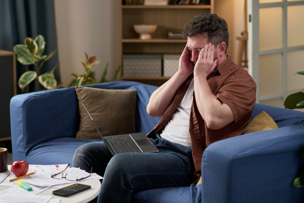 A stressed remote worker on the couch looking at his laptop, with a bunch of papers on his table.